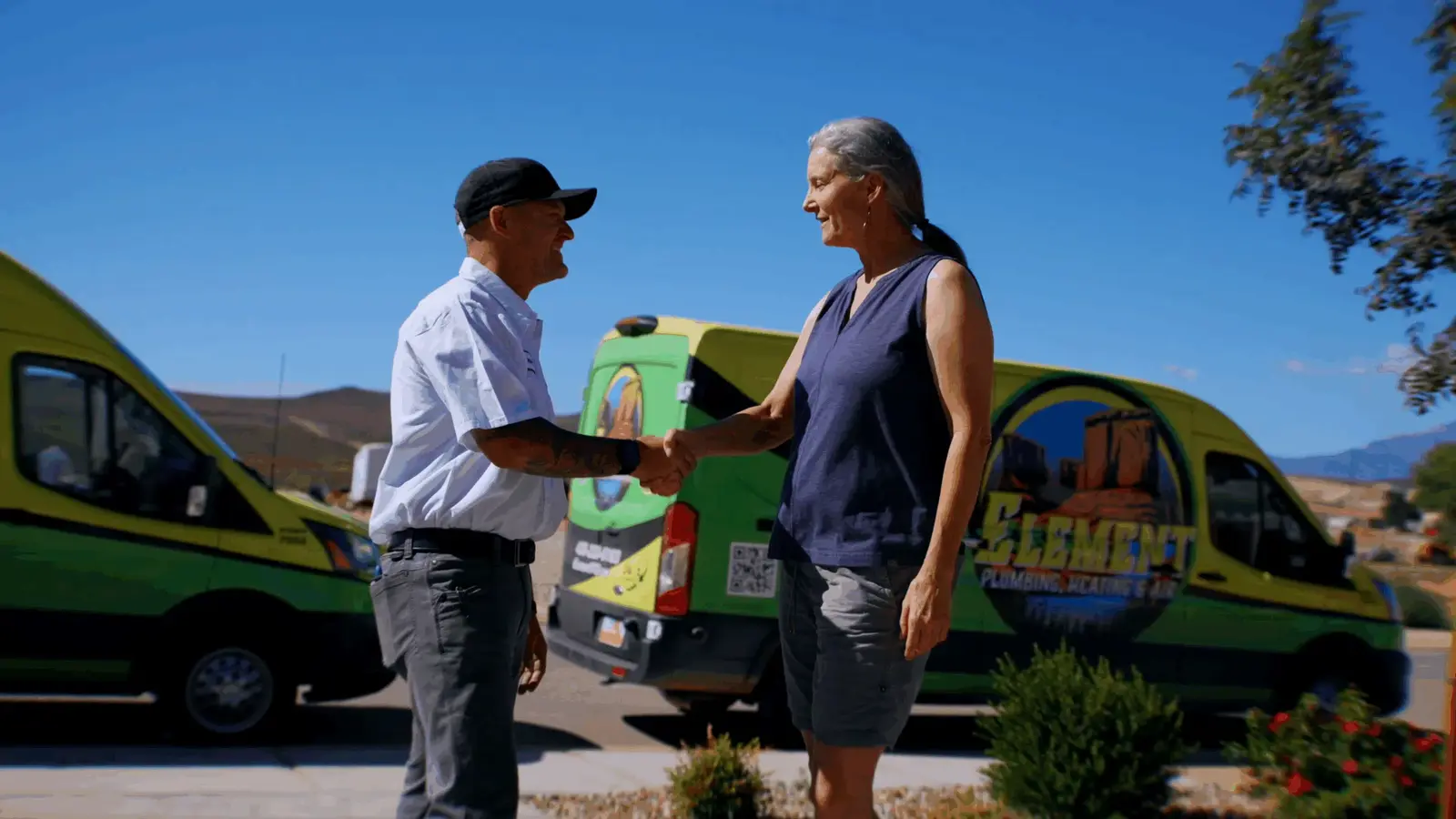Element Plumbing Heating Air technician shaking hands with a customer in St. George UT in front of branded service vans with a scenic mountain backdrop